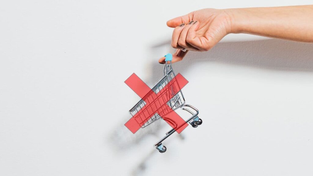 A person's hand holding a small toy shopping cart between their fingers against a white background. A red 'X' is superimposed over the shopping cart, emphasizing the inefficiency that WooCommerce One Page Checkout aims to eliminate.